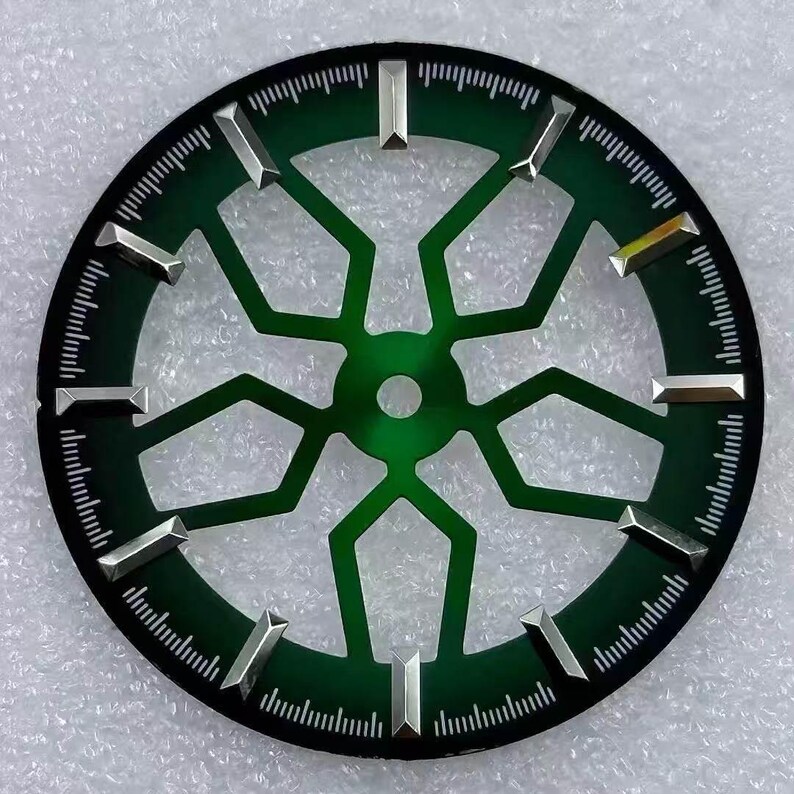 Pode incluir: Um mostrador de rel&oacute;gio com um centro verde escuro e um anel externo preto. O mostrador apresenta marcadores de hora prateados e um design geom&eacute;trico. O anel externo tem pequenas marca&ccedil;&otilde;es brancas para minutos e segundos.