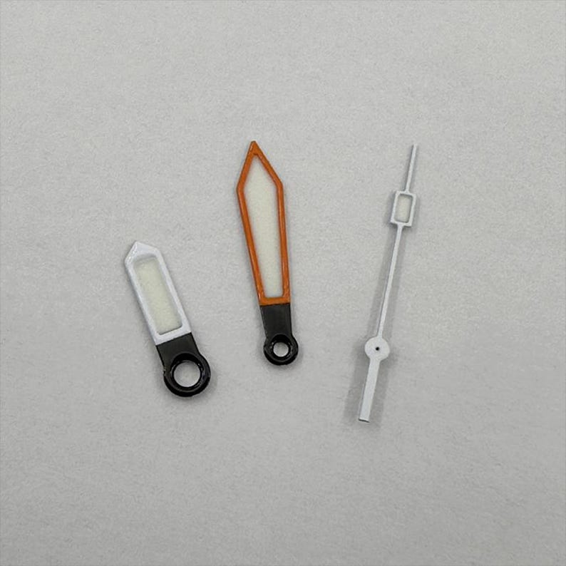 May include: Three watch hands on a gray surface. Two hands have a black base with a white and orange design. The third hand is white with a rectangular tip. The hands are for a watch.