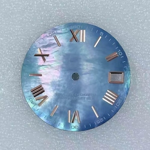 Könnte beinhalten: Ein Zifferblatt mit einer perlmuttartigen blauen Oberfläche, mit römischen Ziffern und Markierungen in Roségold. Das Zifferblatt hat ein kreisförmiges Design mit einer Minutenspur am Rand und dem Text "SUPERLATIVE CHRONOMETER OFFICIALLY CERTIFIED".