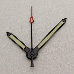 Puede incluir: Un juego de agujas de reloj, que incluye las agujas de las horas, los minutos y los segundos. Las agujas de las horas y los minutos son negras con un relleno verde luminoso, mientras que la aguja de los segundos es roja.