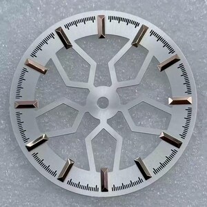 Pode incluir: Um mostrador de rel&oacute;gio prateado com um design geom&eacute;trico e marcadores de hora retangulares dourados. O mostrador tem uma borda externa circular com pequenas marca&ccedil;&otilde;es pretas para os minutos. O centro apresenta um padr&atilde;o vazado estilizado.