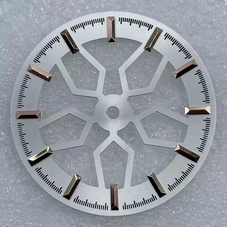 Pode incluir: Um mostrador de rel&oacute;gio prateado com um design geom&eacute;trico e marcadores de hora retangulares dourados. O mostrador tem uma borda externa circular com pequenas marca&ccedil;&otilde;es pretas para os minutos. O centro apresenta um padr&atilde;o vazado estilizado.