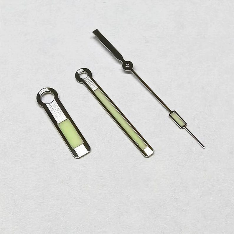 May include: Three watch hands: a short, rectangular one with a circular end, a longer, straight one, and a thin, pointed one. All have a silver finish with a glowing green center. The hands are designed for a watch.
