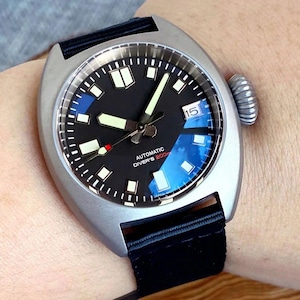 K&ouml;nnte beinhalten: Eine silberfarbene automatische Taucheruhr mit schwarzem Zifferblatt, Leuchtzeigern und Datumsanzeige. Die Uhr hat ein schwarzes Nylonarmband und eine strukturierte Krone. Das Zifferblatt hat blaue Akzente und den Text "AUTOMATIC DIVER'S 200m".