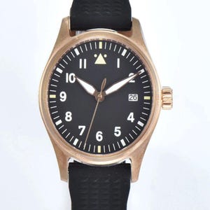 May include: A bronze-colored wristwatch with a black face and white numerals. The watch has a black rubber band and a date display at the 3 o'clock position. The watch hands are white and the second hand is bronze.