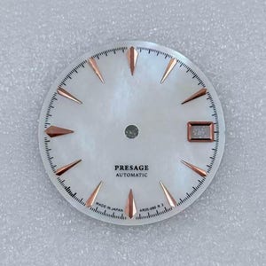 Puede incluir: Una esfera de reloj con fondo blanco nacarado, con marcadores y agujas de color oro rosa. La esfera tiene el texto "PRESAGE AUTOMATIC" y "MADE IN JAPAN". La esfera del reloj tiene una ventana de fecha y un peque&ntilde;o dial de segundos.