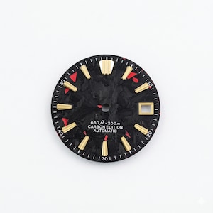 Könnte beinhalten: Ein Zifferblatt mit einem schwarzen Kohlefaser-Muster, goldfarbenen Stundenmarkierungen und roten Akzenten. Der Text "660 ft = 200m CARBON EDITION AUTOMATIC" ist auf dem Zifferblatt aufgedruckt. Das Zifferblatt hat eine runde Form.