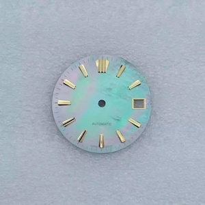 May include: A watch face with a pearlescent, teal-green background. Gold-colored hour markers and hands are visible. The word "AUTOMATIC" is printed at the bottom. The watch face has a square window at the 3 o'clock position.