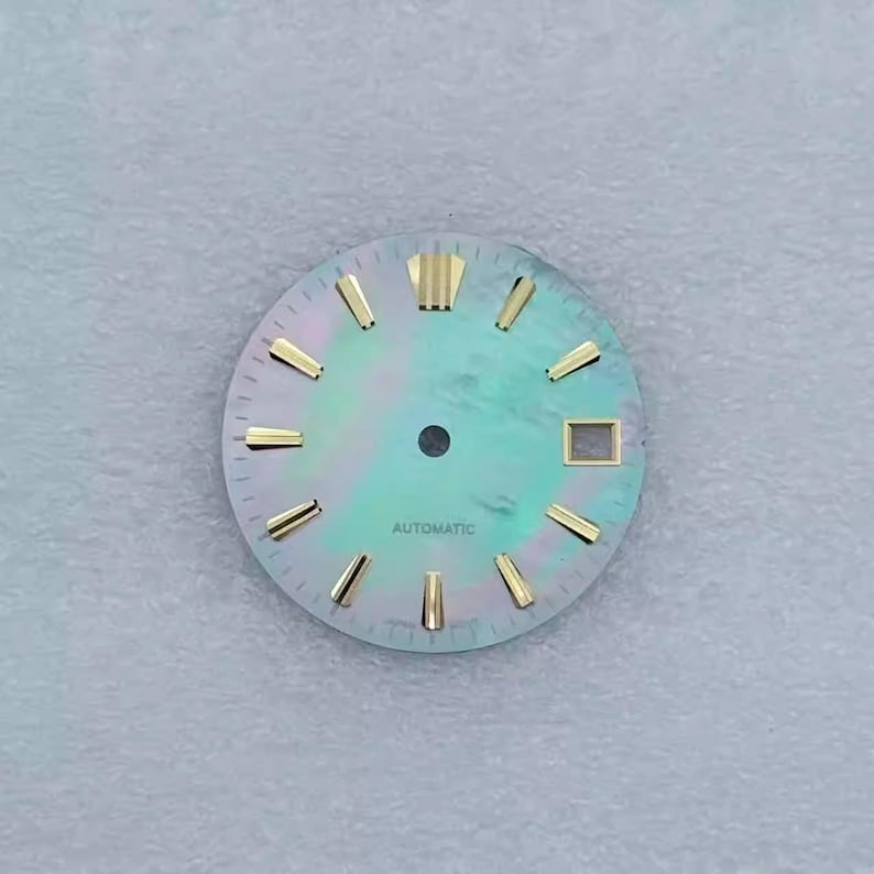May include: A watch face with a pearlescent, teal-green background. Gold-colored hour markers and hands are visible. The word "AUTOMATIC" is printed at the bottom. The watch face has a square window at the 3 o'clock position.