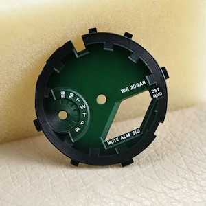 May include: A circular watch dial component featuring a deep green center with a black outer ring. The dial includes white printed text reading WR 20BAR, DST, MND, and MUTE ALM SIG, along with a sub-dial displaying days of the week from SU to S.