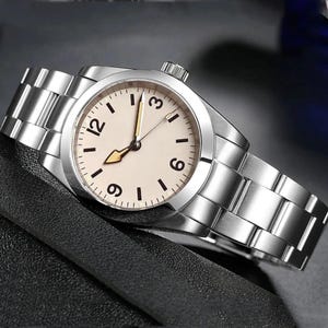 May include: A silver wristwatch with a cream-colored face and black hour markers. The watch has a silver band and a crown on the side. The hands are gold-colored. The watch is displayed on a black surface.