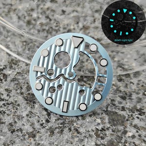 May include: A light blue watch dial with silver accents and a textured surface. The dial features various shapes and circular markers. A smaller image shows the dial glowing in the dark, with the text "BGW9 night light".