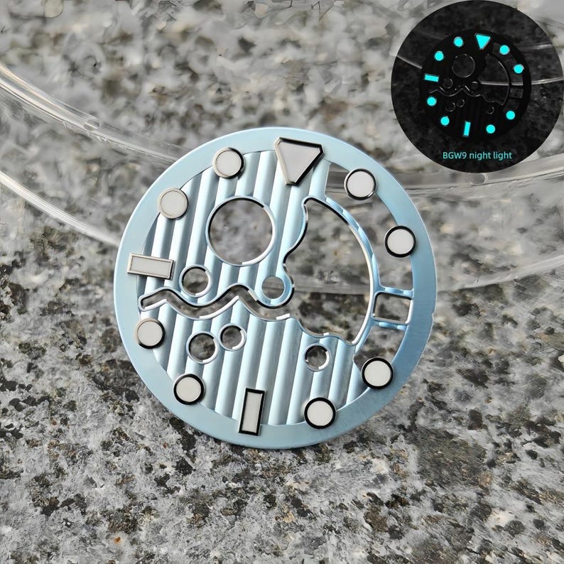 May include: A light blue watch dial with silver accents and a textured surface. The dial features various shapes and circular markers. A smaller image shows the dial glowing in the dark, with the text "BGW9 night light".