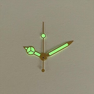 May include: Close-up of watch hands with a gold finish and glowing green luminescence. The hands include a minute hand, hour hand, and second hand. The hands are set against a neutral background.