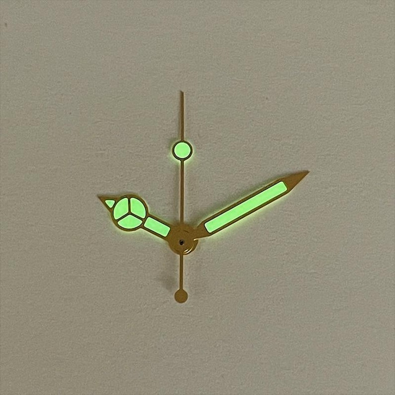 May include: Close-up of watch hands with a gold finish and glowing green luminescence. The hands include a minute hand, hour hand, and second hand. The hands are set against a neutral background.
