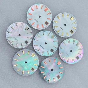 May include: Seven watch dials with iridescent mother-of-pearl faces. Each dial has a central hole, minute markers, and hour markers in various metallic colors. The word "AUTOMATIC" is printed on each dial.