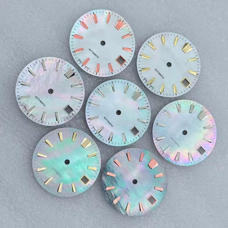 May include: Seven watch dials with iridescent mother-of-pearl faces. Each dial has a central hole, minute markers, and hour markers in various metallic colors. The word "AUTOMATIC" is printed on each dial.