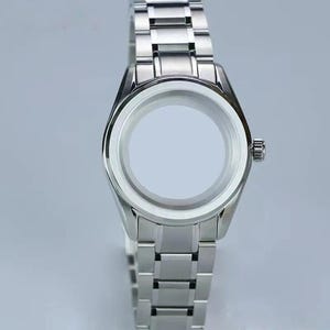 Puede incluir: Una caja de reloj de acero inoxidable en tono plateado con una pulsera de eslabones a juego integrada. La cabeza circular del reloj presenta un bisel pulido y una corona lateral, pero carece de esfera, manecillas o mecanismo interno.