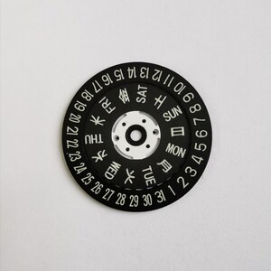 May include: Black watch face with white numbers and day of the week indicators in English and Japanese. The numbers 1 through 31 are displayed around the outer edge of the face, and the days of the week are displayed in a circle in the center. The days of the week are displayed in English and Japanese.