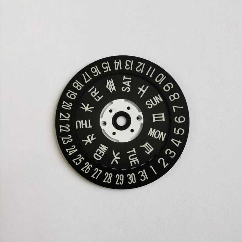 May include: Black watch face with white numbers and day of the week indicators in English and Japanese. The numbers 1 through 31 are displayed around the outer edge of the face, and the days of the week are displayed in a circle in the center. The days of the week are displayed in English and Japanese.