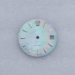 May include: A watch face with a pearlescent, iridescent finish. The face has silver-colored hour markers and hands, with the word "AUTOMATIC" printed in black. The watch face also has a date window and the word "JAPAN" printed at the bottom.