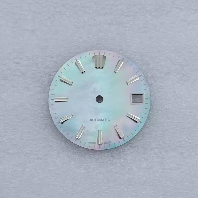 May include: A watch face with a pearlescent, iridescent finish. The face has silver-colored hour markers and hands, with the word "AUTOMATIC" printed in black. The watch face also has a date window and the word "JAPAN" printed at the bottom.