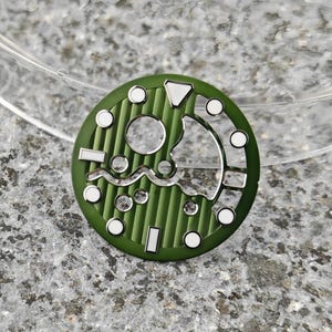 May include: A green watch dial with a textured, striped pattern. The dial features silver-colored cutouts and white circular and rectangular markers. The design includes a wave-like silver accent and a triangular marker.
