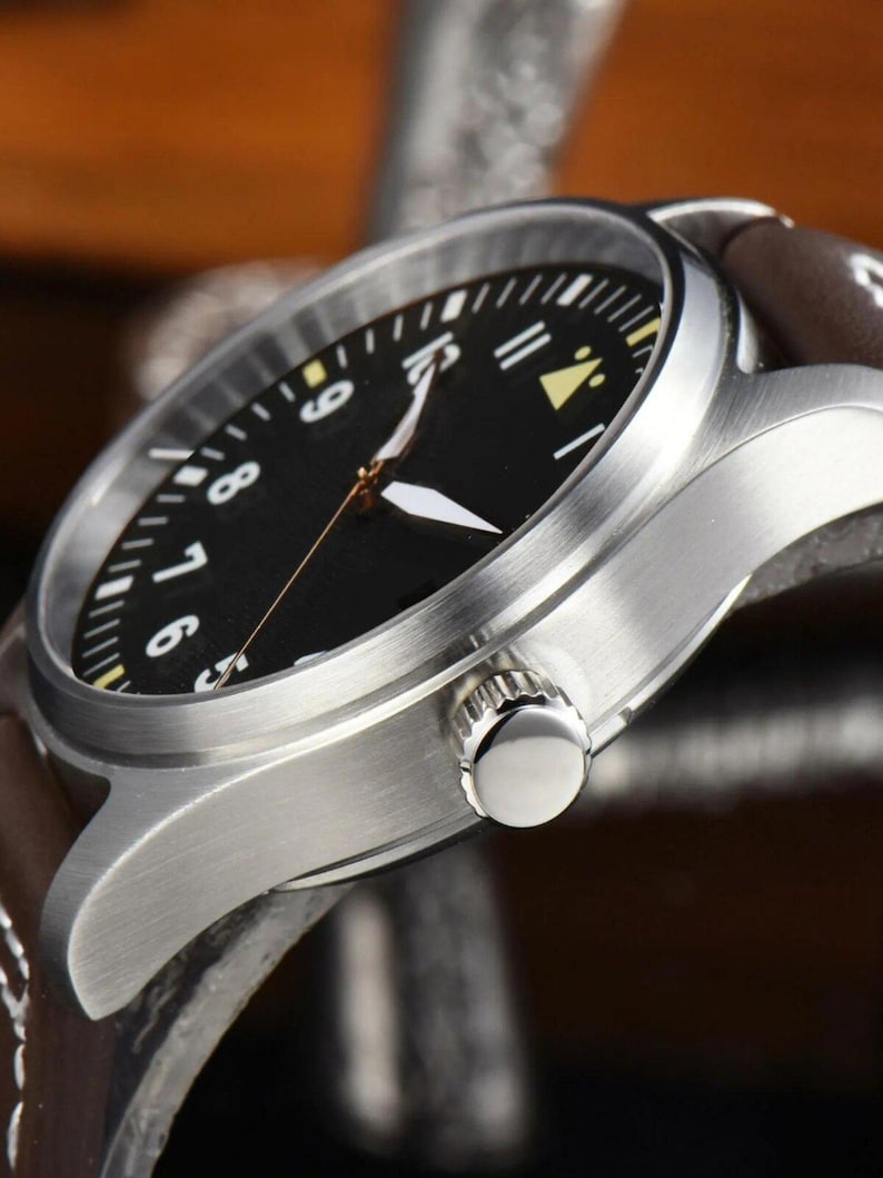 May include: Close-up of a stainless steel wristwatch with a black face and brown leather band. The watch features white and yellow hour markers and hands. The watch's crown is visible on the side.