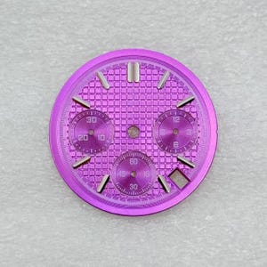 Pode incluir: Um mostrador de relógio circular roxo com textura de grade, marcadores de hora prateados e três submostradores. Os submostradores exibem marcações numéricas para 30, 20, 10, 12, 9, 6, 3, 60, 45, 30 e 15. Uma pequena janela de data é visível.