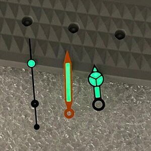 May include: Three watch hands with a glowing green effect. One is a thin black line with a green circle and black dot. Another is orange with a green center. The third is black with a green geometric design.