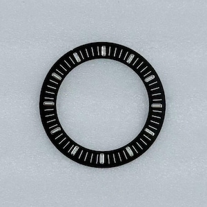 May include: A circular black watch bezel insert featuring a series of white printed minute markers around the perimeter and twelve small, rectangular silver-toned hour indices positioned at regular intervals along the inner edge of the ring.
