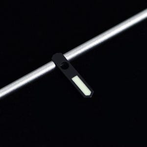 May include: A black watch hand with a white rectangular marker and a silver metal rod on a black background. The watch hand has a small circular hole near the top.