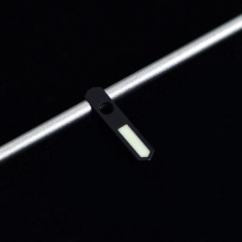 May include: A black watch hand with a white rectangular marker and a silver metal rod on a black background. The watch hand has a small circular hole near the top.