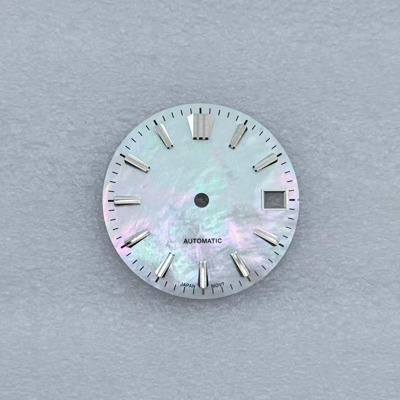 May include: A watch face with a pearlescent, iridescent finish. The face features silver-toned hour markers and hands, with the word "AUTOMATIC" printed in black. The bottom of the face reads "JAPAN MOVT".