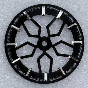 Pode incluir: Um mostrador de rel&oacute;gio preto com um design geom&eacute;trico. O mostrador apresenta marcadores de hora prateados e uma s&eacute;rie de linhas ao redor da borda externa. O centro do mostrador tem uma abertura circular.