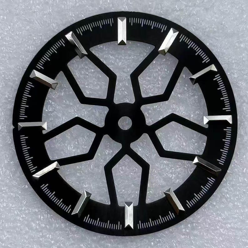 Pode incluir: Um mostrador de rel&oacute;gio preto com um design geom&eacute;trico. O mostrador apresenta marcadores de hora prateados e uma s&eacute;rie de linhas ao redor da borda externa. O centro do mostrador tem uma abertura circular.