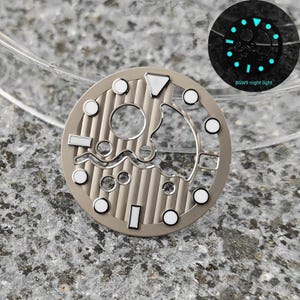 May include: A watch dial with a beige finish, featuring a series of circular and rectangular markers with white accents. The dial has a unique open design with a textured background. A smaller inset shows the dial glowing with a blue night light. The text "BGW9 night light" is visible.
