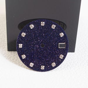 May include: A circular watch dial featuring a deep blue, glittery surface that resembles a starry night sky. It includes twelve square-shaped hour markers with embedded clear stones and a rectangular date window positioned at the three o'clock mark.