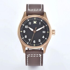 May include: A bronze-colored wristwatch with a black face and white numerals. The watch has a brown leather band with white stitching. The date is displayed at the 3 o'clock position. The watch has a classic, pilot-style design.