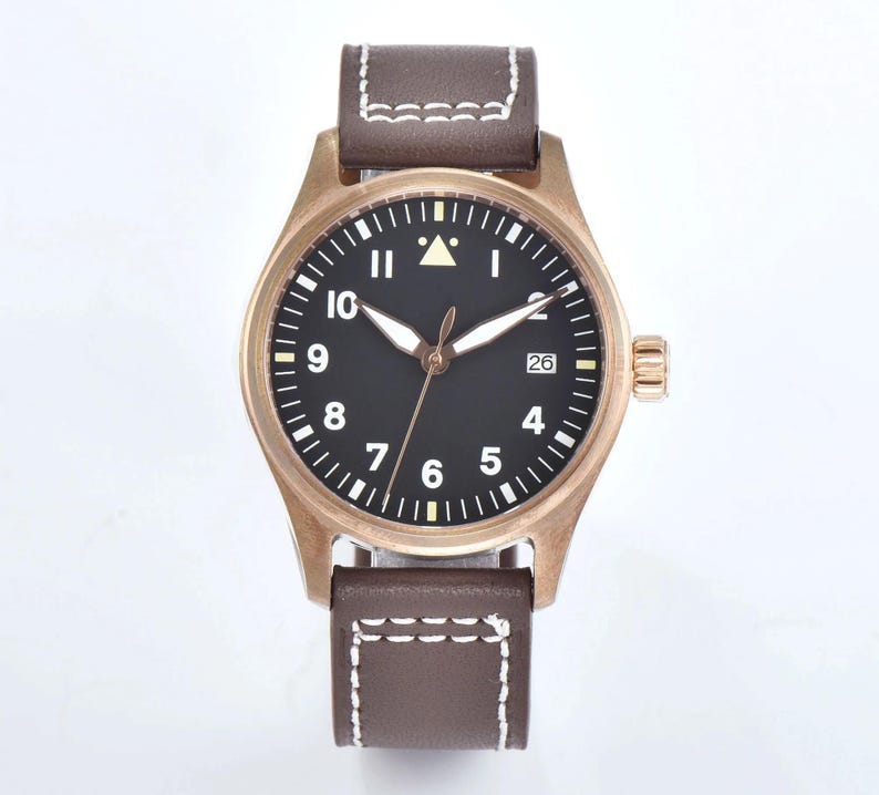 May include: A bronze-colored wristwatch with a black face and white numerals. The watch has a brown leather band with white stitching. The date is displayed at the 3 o'clock position. The watch has a classic, pilot-style design.