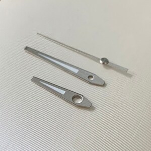 Pu&ograve; includere: Tre lancette di orologio argentate con accenti bianchi. Le lancette sono per un orologio con un quadrante analogico tradizionale.