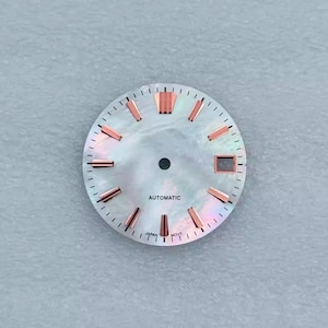 May include: A watch face with a pearlescent, iridescent finish. It features rose gold-colored hour markers and a small date window. The word "AUTOMATIC" is printed at the center, with "JAPAN MOVT" at the bottom.