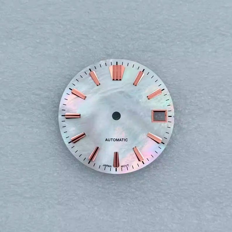 May include: A watch face with a pearlescent, iridescent finish. It features rose gold-colored hour markers and a small date window. The word "AUTOMATIC" is printed at the center, with "JAPAN MOVT" at the bottom.