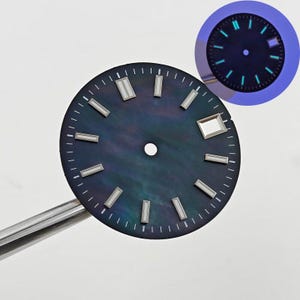Può includere: Un quadrante di orologio con una superficie scura e iridescente e indici delle ore rettangolari argentati. Il quadrante ha una piccola finestra della data ed è tenuto da uno strumento d'argento. Una versione più piccola e illuminata del quadrante mostra indicatori verdi luminosi.