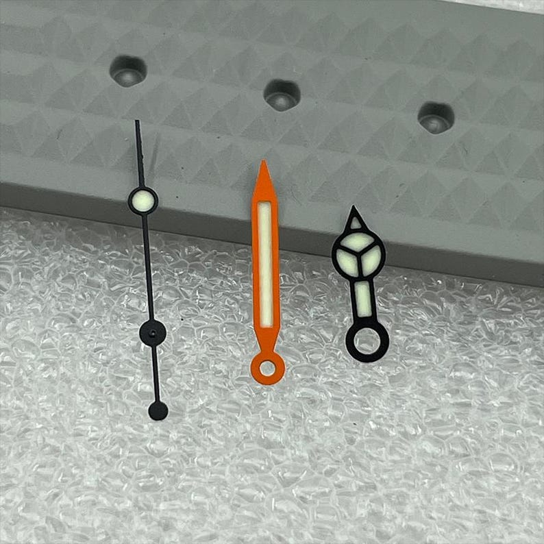 May include: Three watch hands in a row. The first is black with a white circle and a black circle. The second is orange with a white stripe. The third is black with a white triangle and a white circle.