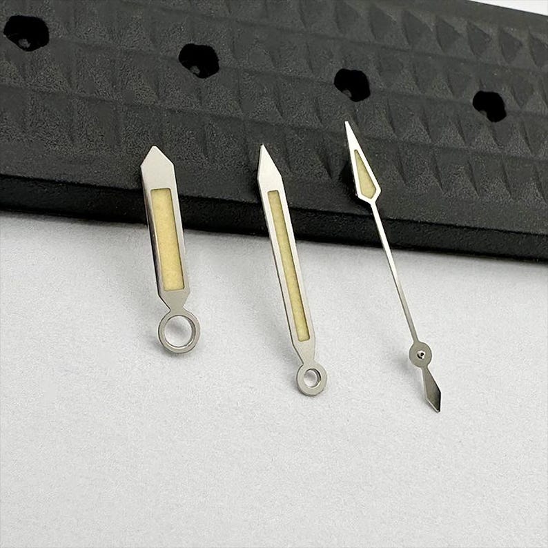 May include: Three silver watch hands with luminous yellow-beige centers. The hands have different shapes, including a rectangular design with a circular base and a pointed arrow design. The hands are set against a white surface and a black textured background.