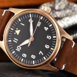 May include: A bronze-colored wristwatch with a black face, white numerals, and luminous hands. The watch has a brown leather strap with white stitching. The watch face displays the date and is set against a dark background.