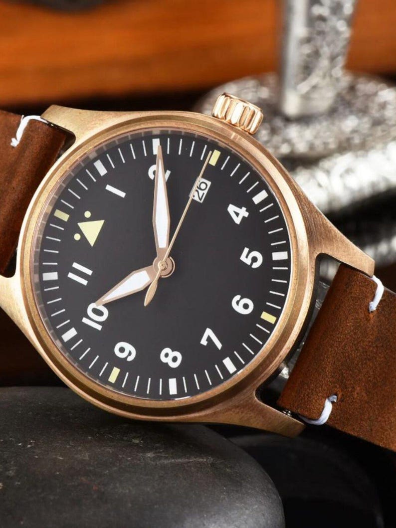 May include: A bronze-colored wristwatch with a black face, white numerals, and luminous hands. The watch has a brown leather strap with white stitching. The watch face displays the date and is set against a dark background.