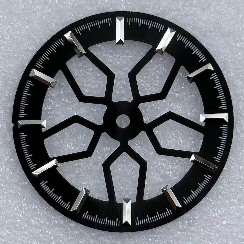 Pode incluir: Um mostrador de rel&oacute;gio preto com um design geom&eacute;trico. O mostrador apresenta marcadores de hora prateados e uma s&eacute;rie de linhas ao redor da borda externa. O centro do mostrador tem uma abertura circular.