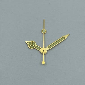 May include: Gold-toned watch hands set against a gray background. The hands include a minute hand with a circular tip, an hour hand with a stylized design, and a second hand with a pointed tip. The hands have a luminous green fill.